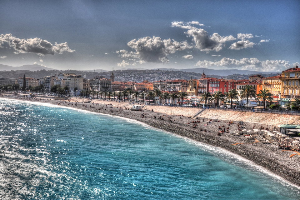 Aerial view of Nice's Promenade des Anglais curving along a turquoise Mediterranean bay, with terracotta rooftops of the old town in the background