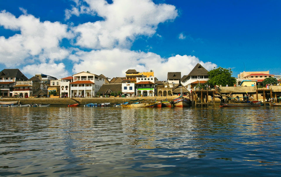 Traditional dhow sailing in Lamu waters with historic stone buildings in background. Photo by Stanislav Ivovsky.